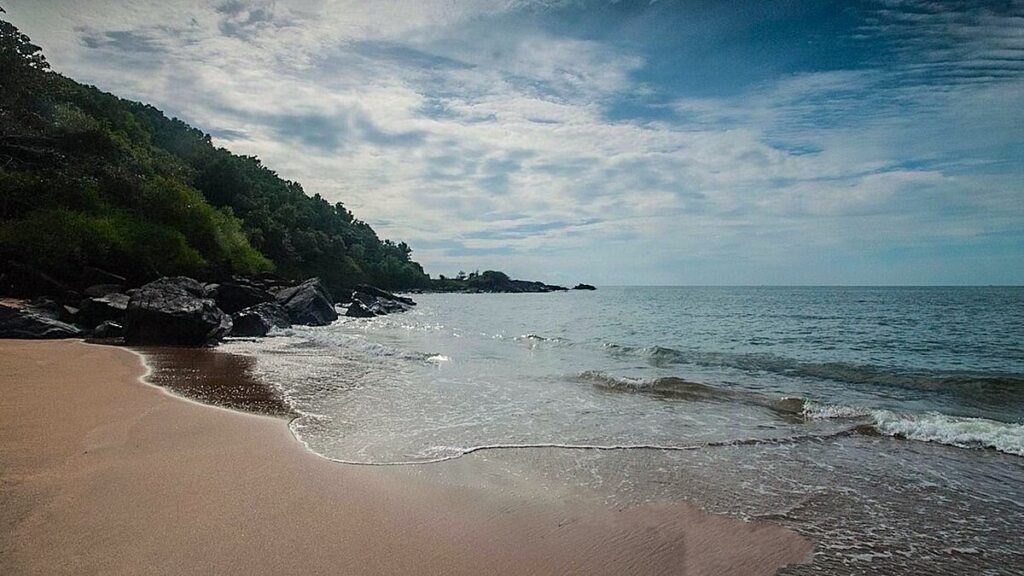 Serene view of Half Moon Beach in Gokarna, Karnataka with soft waves, rocky coastline, and lush greenery under a partly cloudy sky