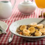 A bowl of banana and cereal served as healthy snacks for kids, placed on a red checkered tablecloth with orange juice in the background.