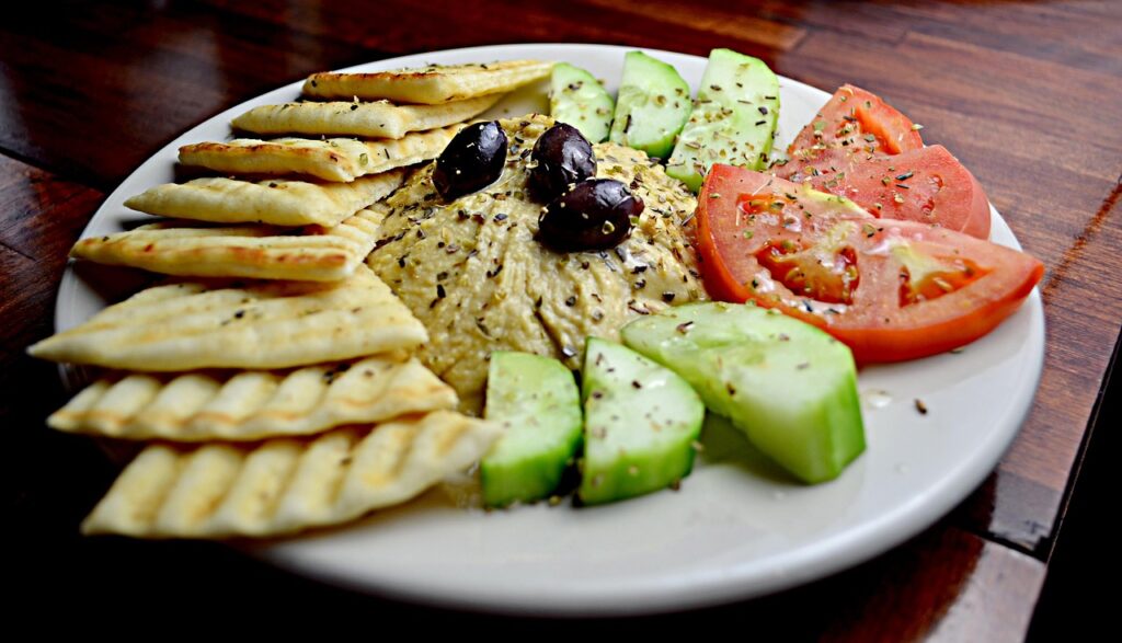 A plate of hummus served with pita crackers, cucumber slices, tomato wedges, and olives as healthy snacks for kids.