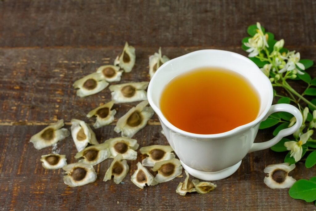 White cup of moringa herbal tea on wooden table with dried moringa seeds and fresh flowers