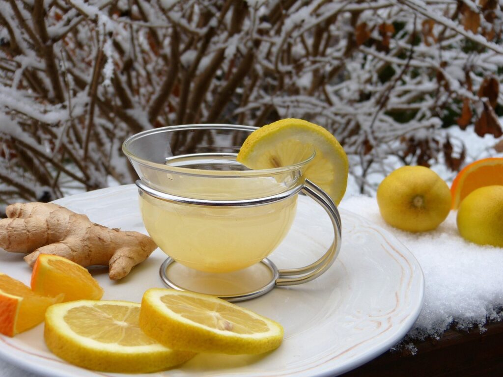 A glass of lemon ginger tea surrounded by fresh lemon slices, ginger root, and oranges on a snowy outdoor table