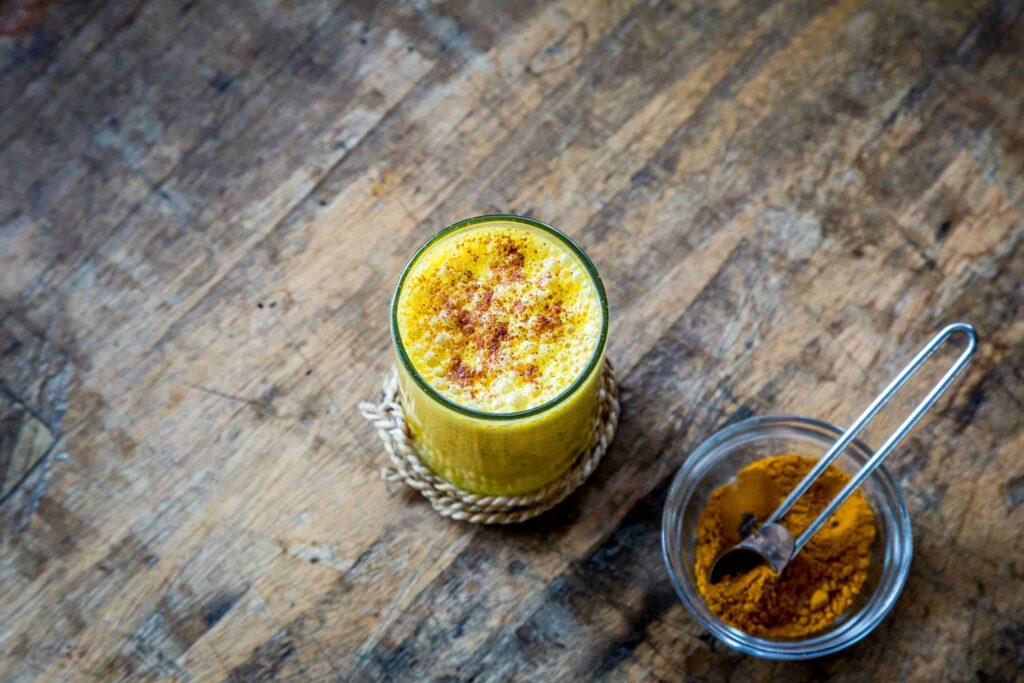 Glass of golden turmeric milk topped with cinnamon, next to a bowl of turmeric powder on a rustic wooden table