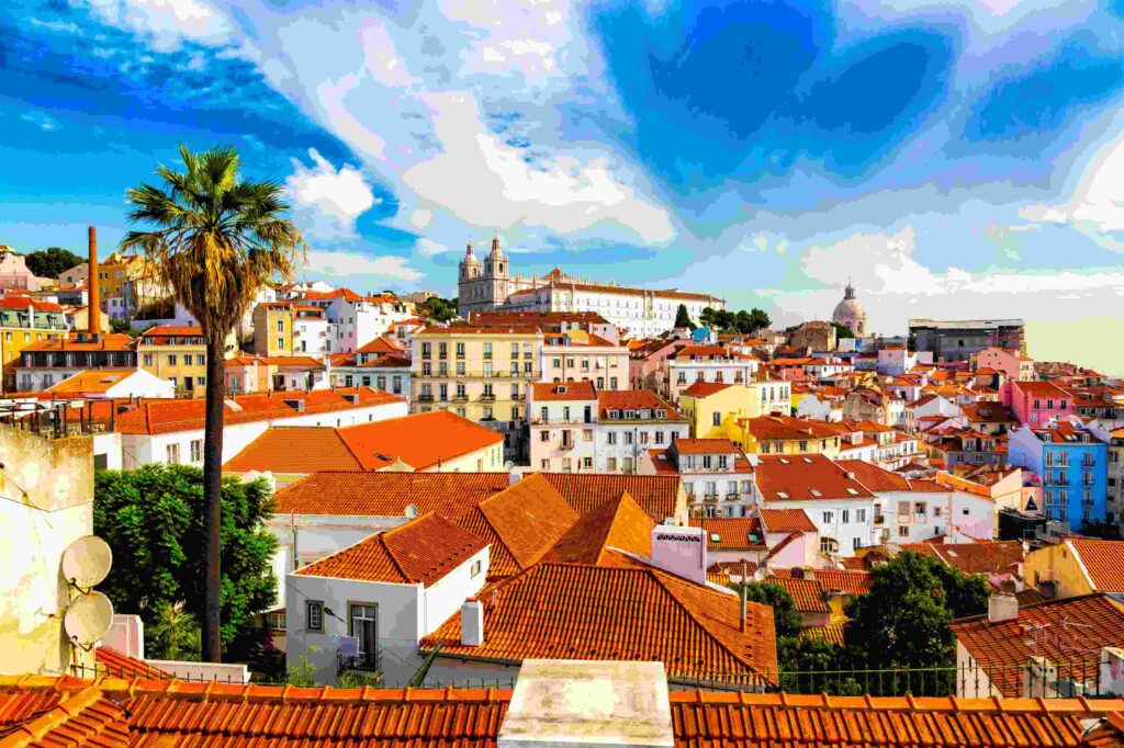 Vibrant cityscape of Lisbon, Portugal, showcasing red-tiled rooftops, pastel-colored buildings, and the historic São Vicente de Fora Monastery under a bright blue sky.