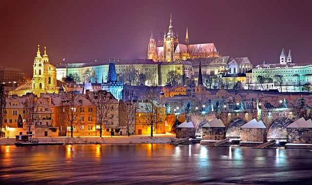Illuminated view of Prague Castle and surrounding historic buildings at night, reflecting beautifully on the Vltava River in winter.