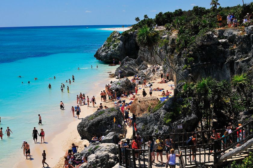 Tourists enjoying the turquoise waters and sandy shores of Tulum, Mexico, with scenic rocky cliffs and palm trees, showcasing it as a budget-friendly honeymoon destination.