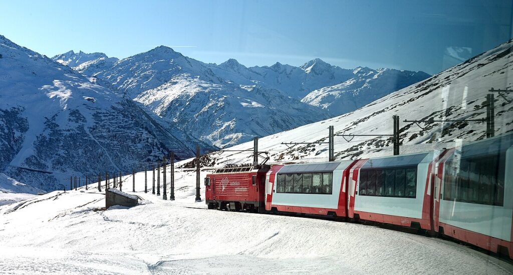 The Glacier Express in Switzerland, one of the most scenic train rides in the world, traveling through the Swiss Alps with snow-covered peaks and lush green valleys in the background.